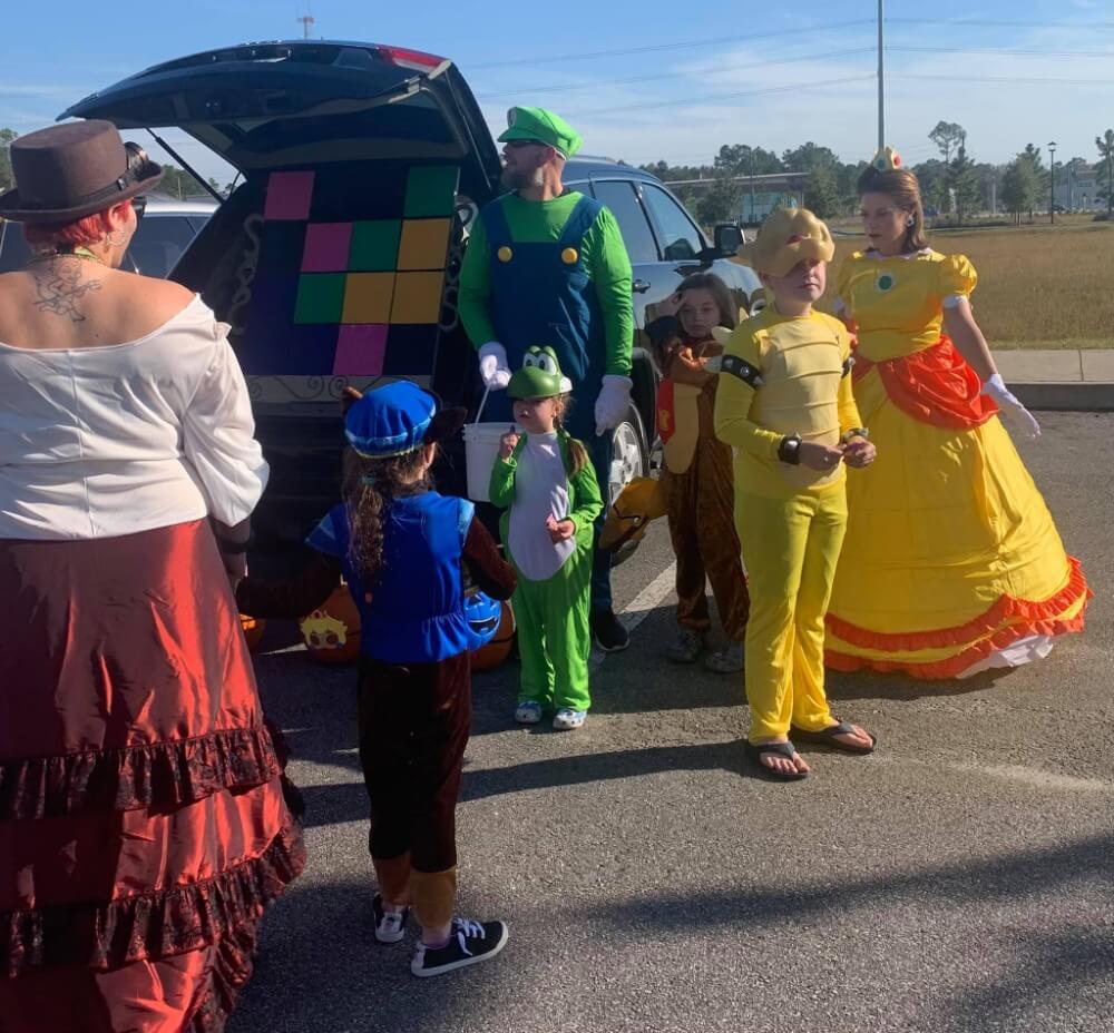 A group of people and children in Mario-themed costumes stand by a car with its trunk open in the sunny parking lot of Wildlight Florida, adding fun to the vibrant atmosphere found near homes in Nassau County Florida.