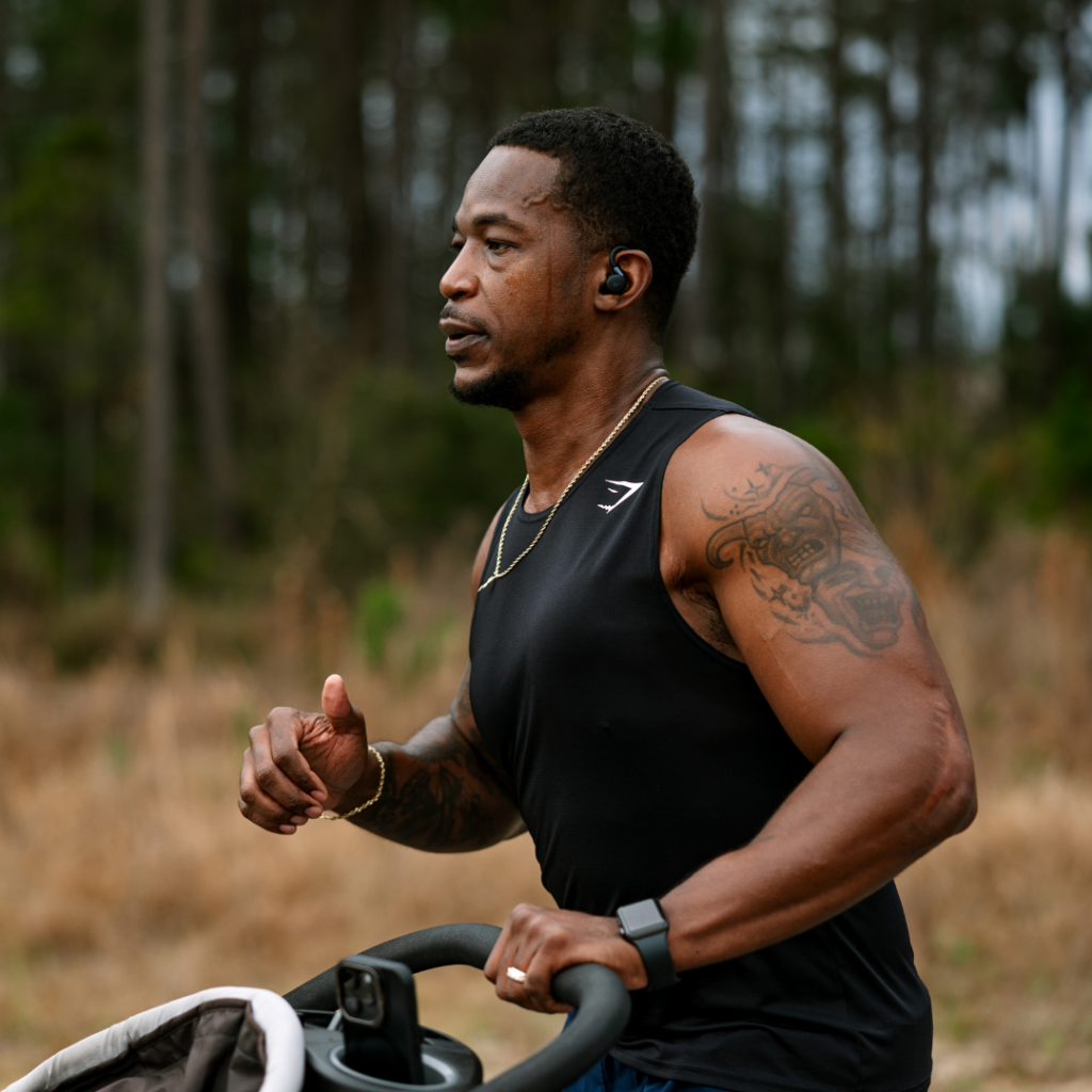 A man in athletic clothing jogs outdoors through the scenic Wildlight Yulee Florida community, pushing a stroller, wearing wireless earbuds and a smartwatch, with trees in the background.