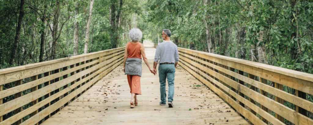 An older couple holds hands while walking on a wooden boardwalk through a green, wooded area in the Wildlight Yulee Florida community.