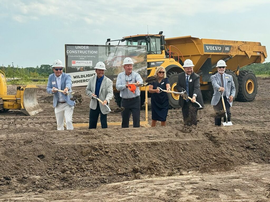 Six people in business attire and hard hats participate in a groundbreaking ceremony with shovels at a construction site, marking the beginning of new homes in Nassau County Florida, with heavy machinery and a project sign in the background.
