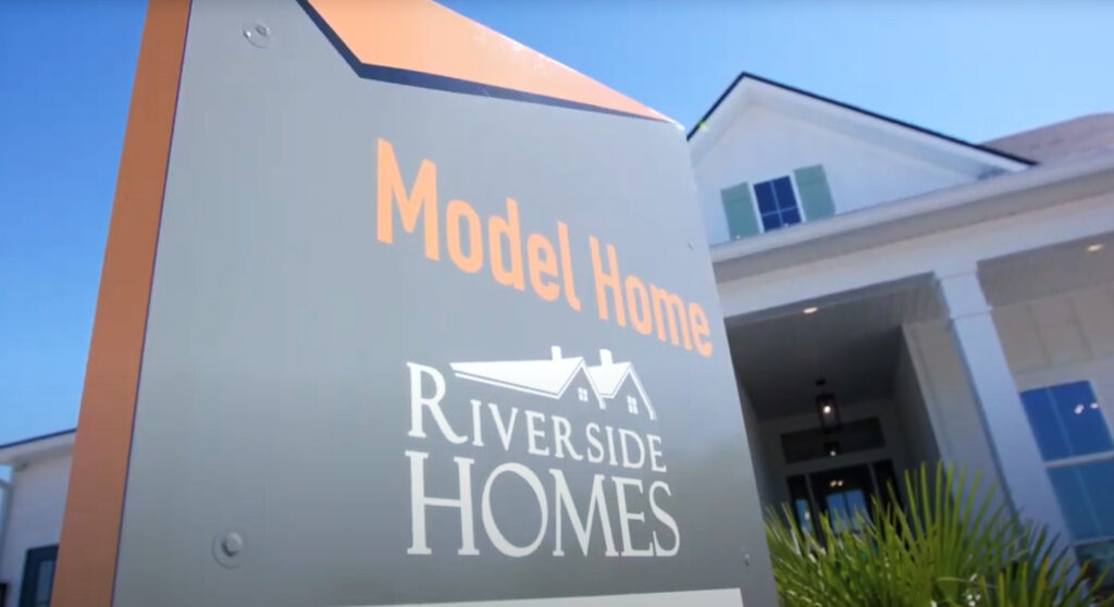 Sign reading "Model Home Riverside Homes" stands in front of a modern white house with columns and green shutters under a clear blue sky, showcasing new homes in Wildlight FL.