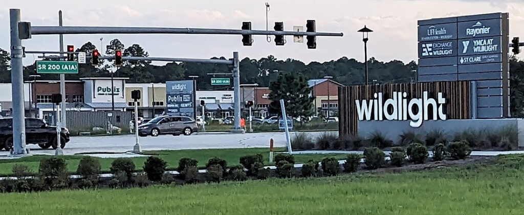 A commercial area with a "Wildlight" entrance sign, traffic lights, and shops like Publix and VyStar along SR 200, set in the Wildlight master planned community known for new homes in Wildlight FL within a vibrant suburban setting.