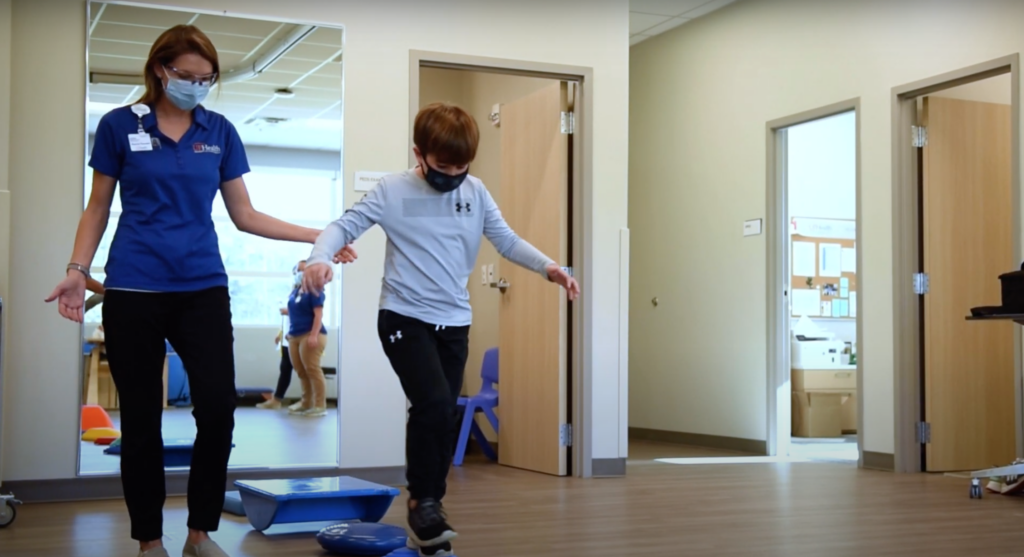 A woman assists a child balancing on one foot in a physical therapy room in the Wildlight Yulee Florida community. Both wear masks, and therapy equipment is visible in the background.