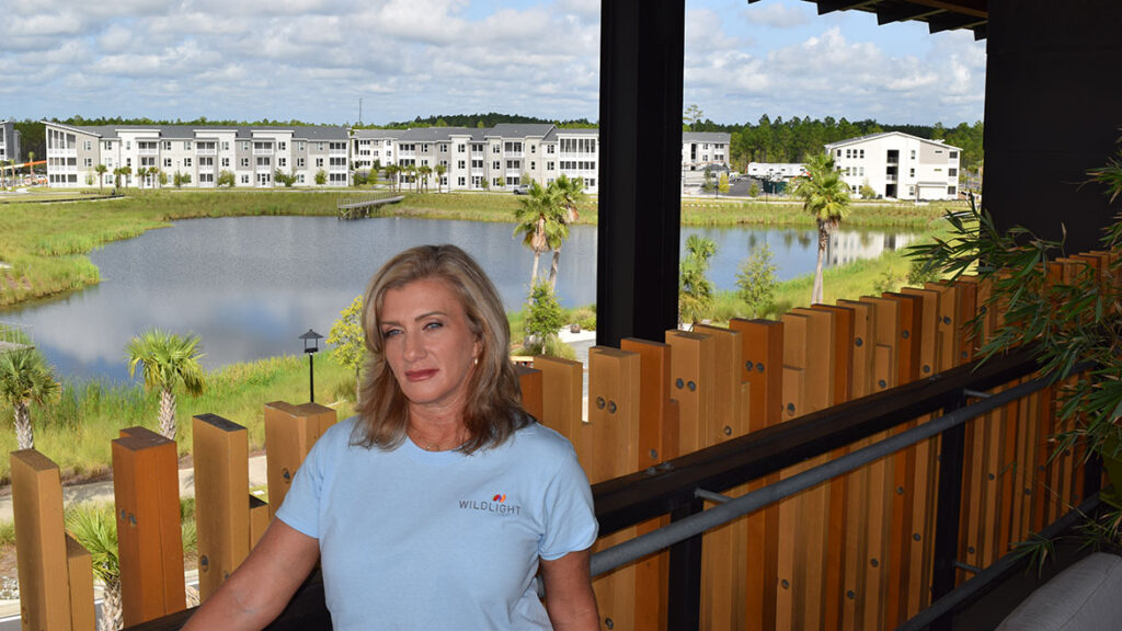 A woman in a light blue shirt stands on a balcony overlooking a pond, grassy area, and modern apartment buildings in the Wildlight master planned community on a partly cloudy day.