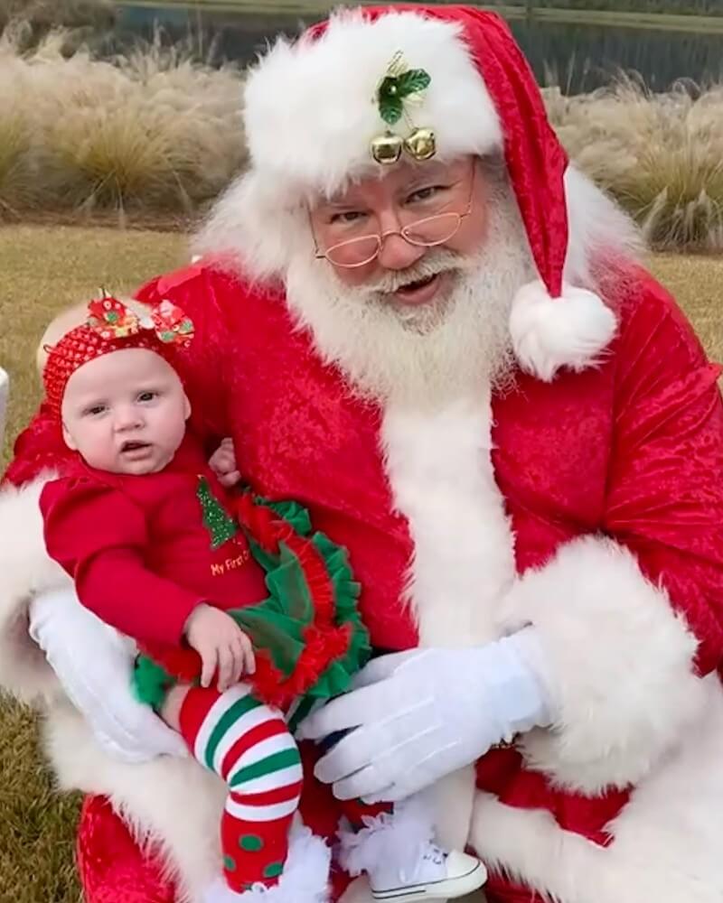 A man dressed as Santa Claus holds a baby in festive red and green holiday clothing while sitting outdoors on the grass, capturing the cheerful spirit of the Wildlight Yulee Florida community.