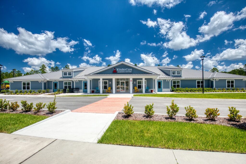 A single-story blue building with a metal roof, labeled “KinderCare Learning Center,” sits under a blue sky with scattered clouds, surrounded by grass and shrubs in the vibrant Wildlight Yulee Florida community.