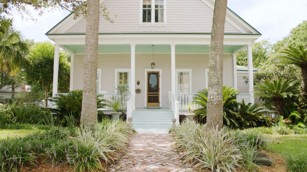 Front view of a light-colored house with a covered porch, two palm trees, and a brick walkway, surrounded by lush greenery—perfect for those seeking homes in Nassau County, Florida's scenic Wildlight master planned community.