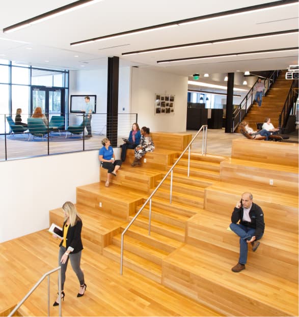 Open office space with wooden tiered seating in the heart of the Wildlight master planned community; several people sitting, talking, and using devices; stairs and glass-walled meeting room in the background.