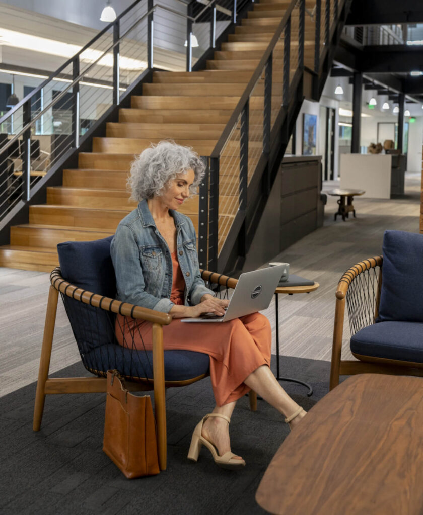 A woman with gray curly hair sits in a modern office lounge in Wildlight Florida, working on a laptop with a brown bag beside her and stairs in the background—perhaps searching for new homes in Wildlight FL.