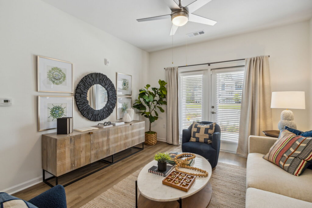 Modern living room in the Wildlight Yulee Florida community, featuring a beige sofa, round coffee table, sideboard with decor, wall art, a large plant, ceiling fan, and glass doors with curtains letting in natural light.