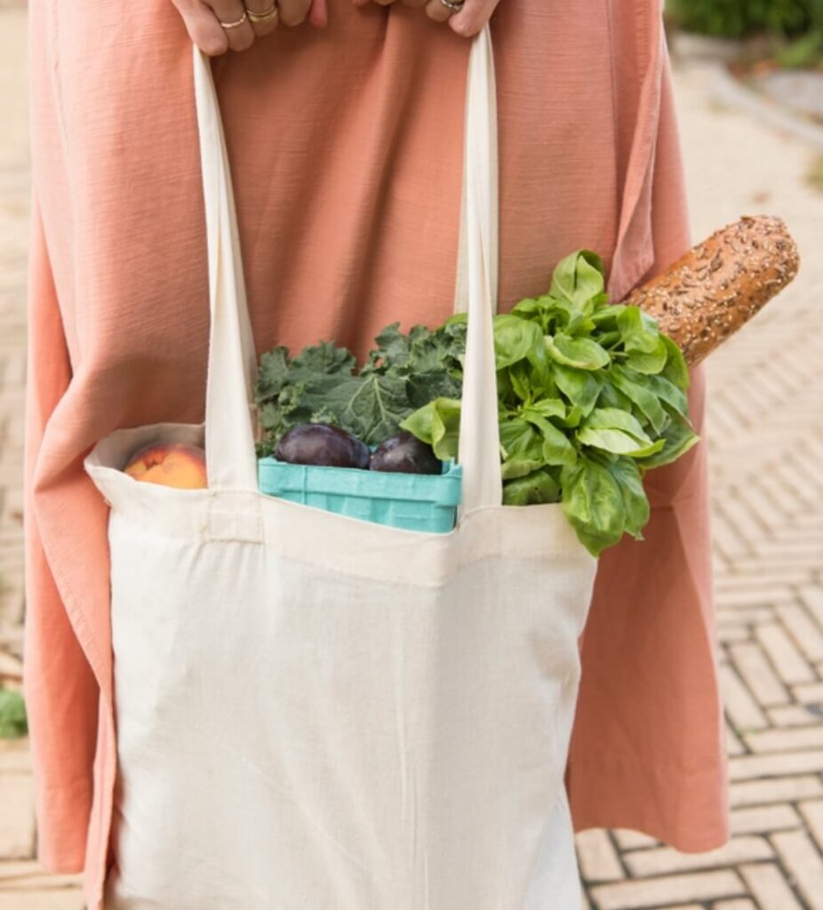 A person holding a tote bag filled with leafy greens, purple produce, a peach, and a multigrain baguette—enjoying fresh finds in the heart of the Wildlight Yulee Florida community.
