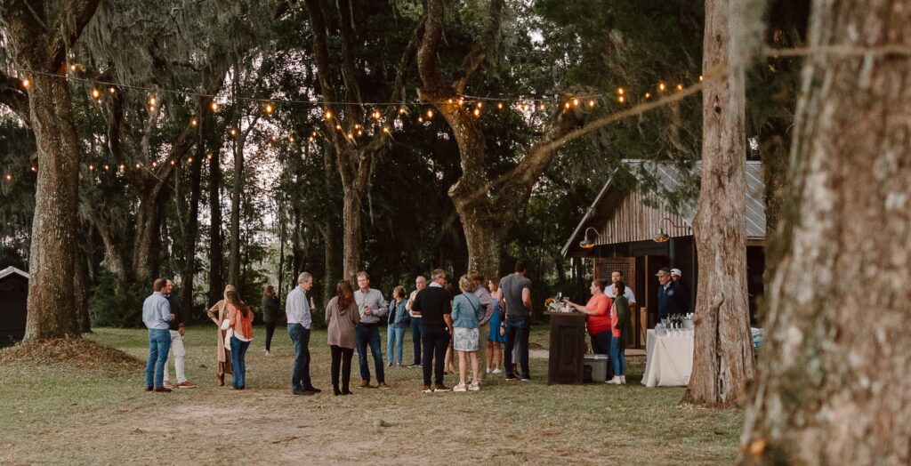 A group of people socialize under string lights in a wooded outdoor area near a rustic wooden building.