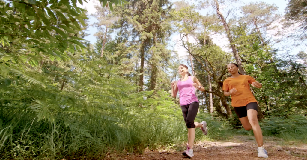 Two women jog along a dirt path through a green, wooded forest on a sunny day in the scenic Wildlight master planned community.