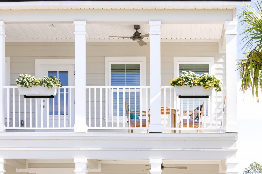 Second-story porch of a white house with planter boxes of flowers, a ceiling fan, wooden table, and chairs—perfect for enjoying bright daylight in the Wildlight master planned community.
