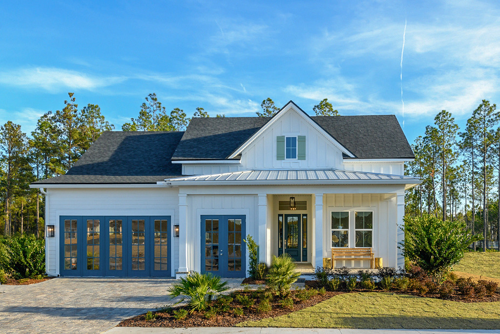 A modern, white single-story house with blue doors, large windows, front porch, and landscaped yard sits in the scenic Wildlight Yulee Florida community, nestled among tall trees beneath a clear sky.