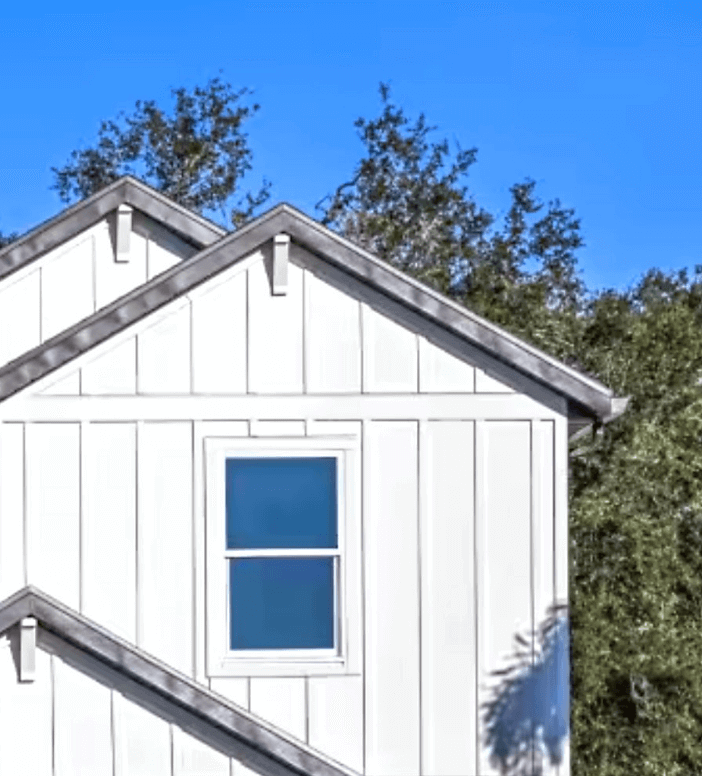 White house exterior with horizontal siding, a single window, and overlapping gable roofs—this charming style is found among new homes in Wildlight FL, where trees frame the landscape under a clear blue sky.