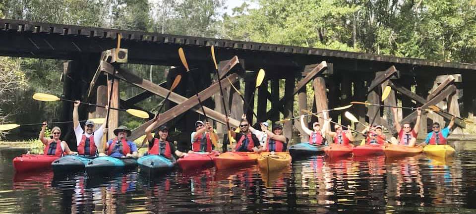 A group of people in colorful kayaks sit side by side on a river in the Wildlight Yulee Florida community, holding up paddles, with a wooden bridge and trees from the scenic Wildlight master planned community in the background.
