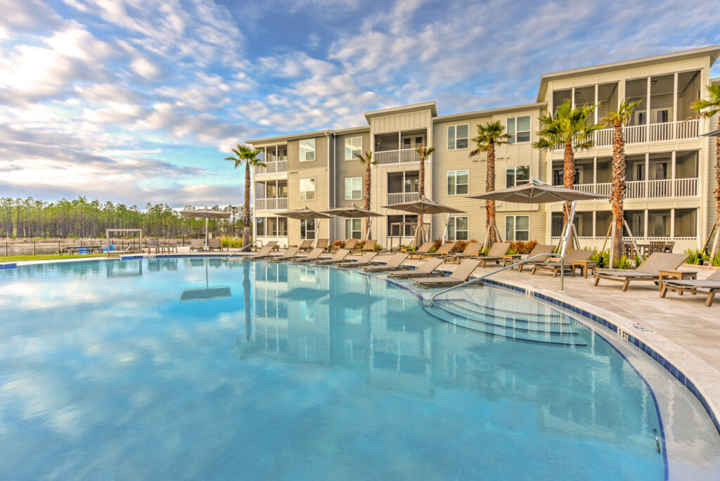 Modern apartment complex with three floors, palm trees, and lounge chairs surrounding a large outdoor swimming pool under a partly cloudy sky in the Wildlight master planned community of Nassau County, Florida.