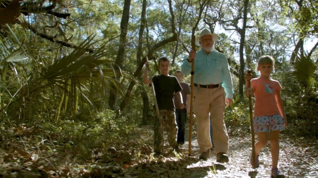 An older man and three children walk with sticks along a sunlit forest trail surrounded by trees and foliage, enjoying the natural beauty that Wildlight Florida offers near new homes in Wildlight FL.