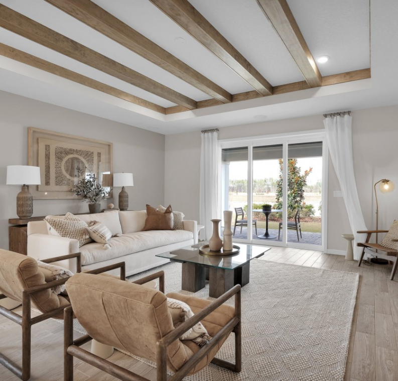 Modern living room with beige furniture, wood ceiling beams, glass coffee table, and large window with white curtains overlooking a patio and green outdoor space in the heart of the Wildlight Yulee Florida community.