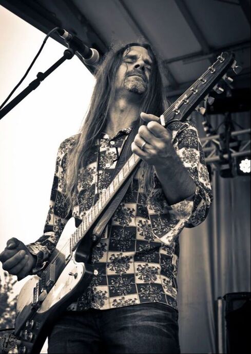 A guitarist with long hair and a patterned shirt plays an electric guitar on stage, entertaining the Wildlight Yulee Florida community as he stands in front of a microphone.