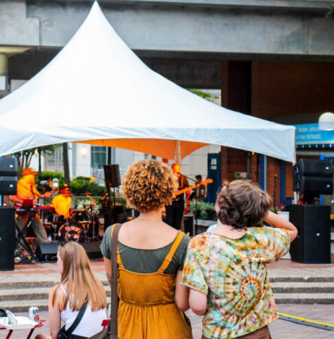 Three people watch a live band perform under a white tent at an outdoor event in the Wildlight Yulee Florida community. Musicians play instruments on stage, bringing lively energy to the vibrant atmosphere.