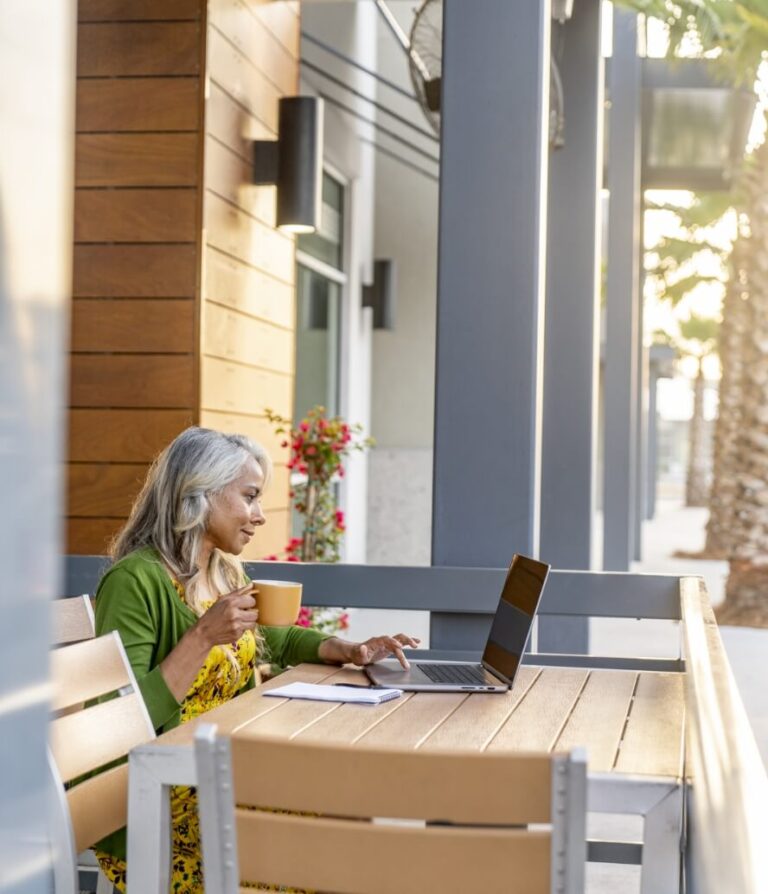 A woman sits at an outdoor table with a laptop and a mug, working and drinking coffee on her patio surrounded by potted plants and palm trees in one of the beautiful new homes in Wildlight FL.