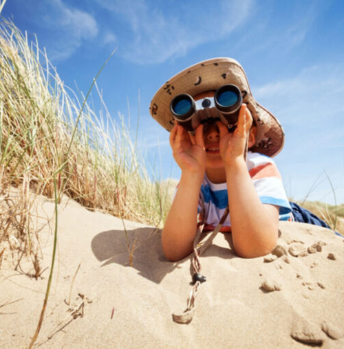 A child in a hat and striped shirt lies on sand among dune grass, looking through binoculars under a blue sky—capturing the wonder you'll find in the Wildlight Yulee Florida community with beautiful new homes in Wildlight FL.