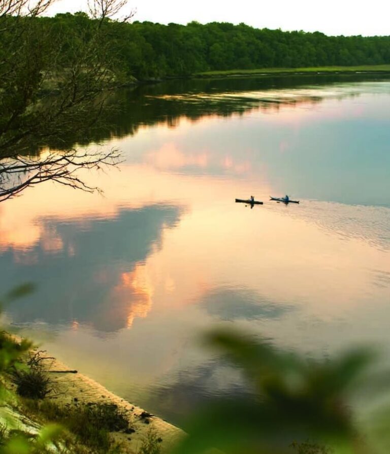 Two people in kayaks paddle on a calm lake at sunset in Wildlight Florida, with trees lining the shore and colorful clouds reflected in the water.