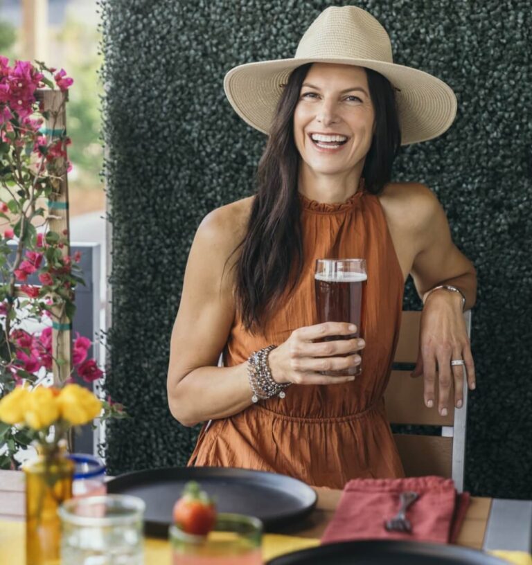 Woman in a sleeveless rust dress and wide-brimmed hat sits at an outdoor table, smiling and holding a glass of dark beverage, enjoying the relaxed vibe of the Wildlight Yulee Florida community surrounded by flowers and plates.