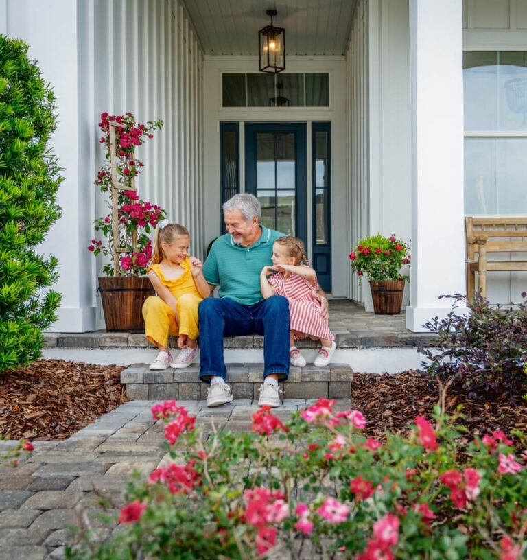 An older man sits on front porch steps with two young girls, surrounded by flowering plants and greenery in front of a white house, reflecting the relaxed lifestyle found in homes in Nassau County Florida.