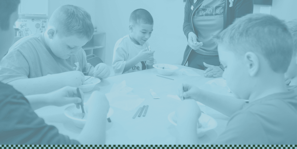 Five children sit around a table eating from bowls, while an adult stands nearby assisting them in a welcoming space within the Wildlight Yulee Florida community.