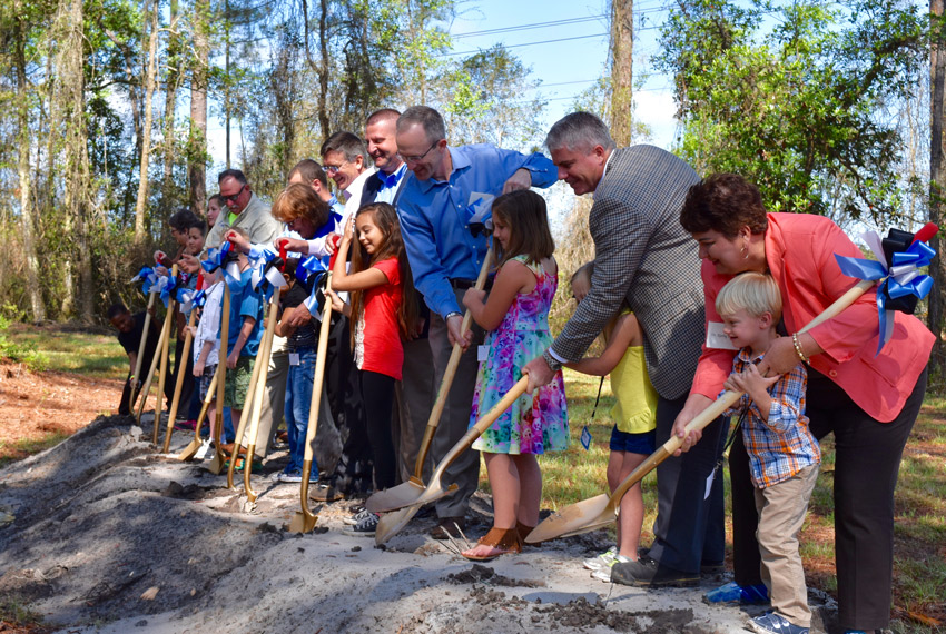 A group of adults and children stand in a line outdoors, each holding a shovel and participating in a ceremonial groundbreaking event for new homes in the Wildlight Yulee Florida community.