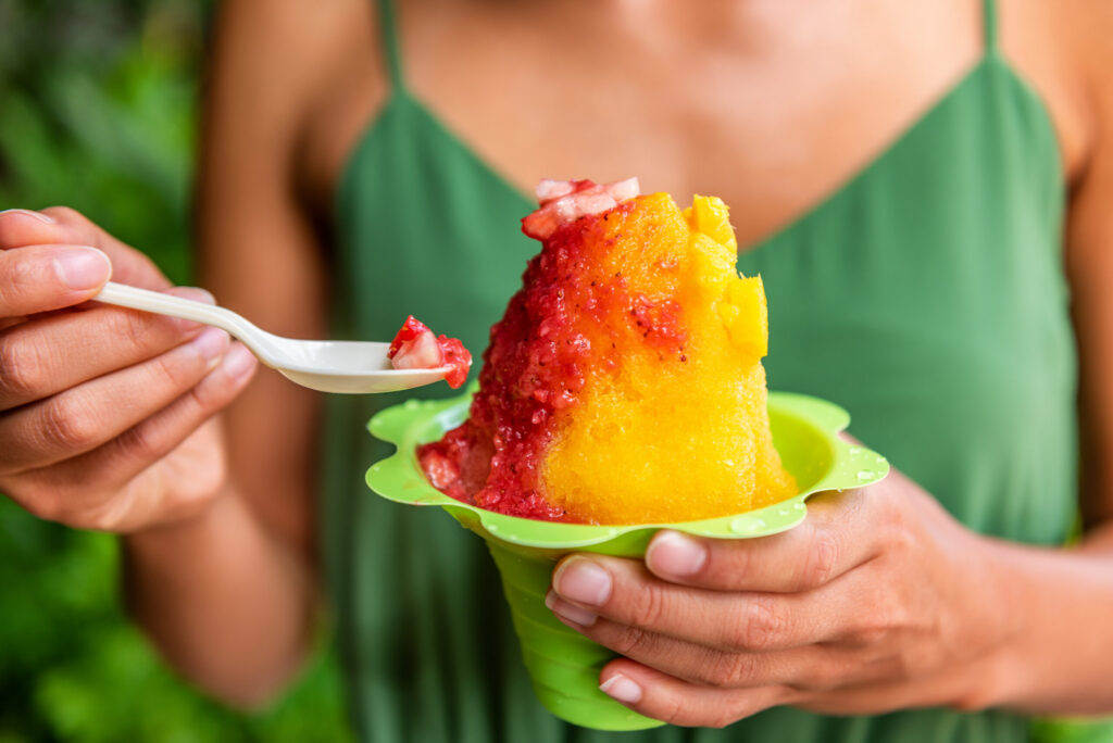 A person in a green top enjoys a yellow and red shaved ice dessert in a green cup, using a spoon to scoop a bite—capturing the sweet moments found in the vibrant Wildlight Yulee Florida community.