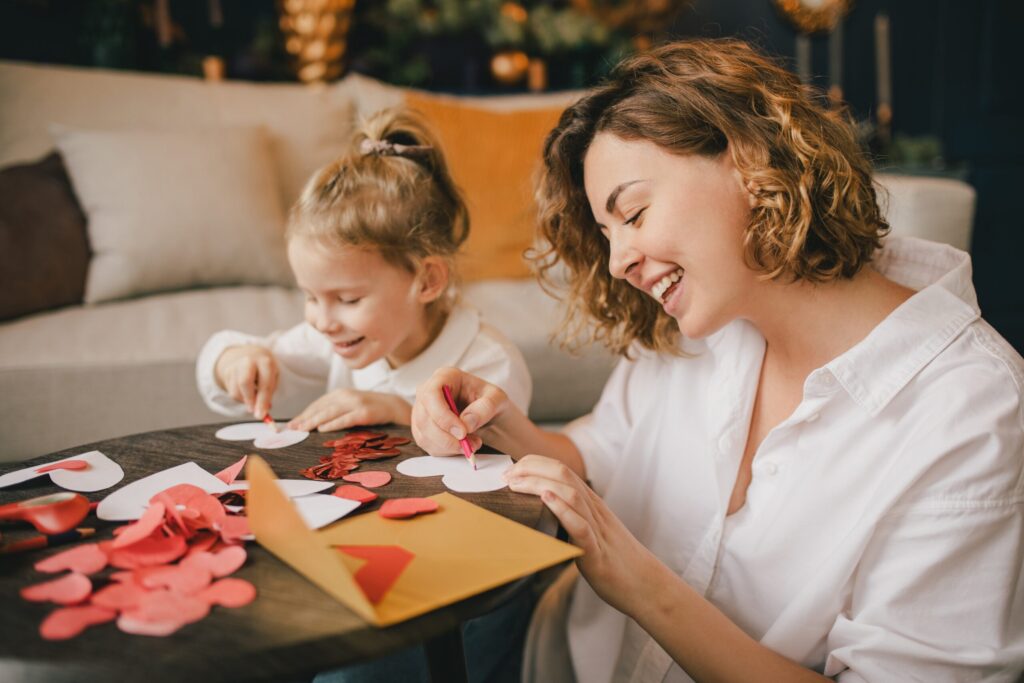 An adult and a child sit at a table making heart-shaped crafts with paper and glitter in their new home in the Wildlight master planned community. The table is covered with craft supplies and an open yellow envelope.