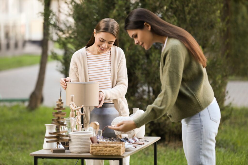 Two women examine items on an outdoor table at a yard sale in the Wildlight Florida community, browsing a lamp, baskets, and decorative objects amid trees and grass in the background.