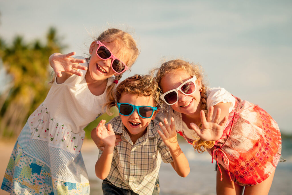 Three smiling children wearing sunglasses wave at the camera on a beach near palm trees and the ocean, embodying the fun and vibrant spirit of the Wildlight Yulee Florida community.