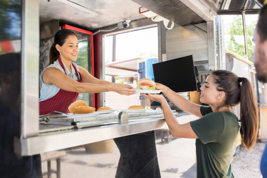 A woman in an apron hands a burger to a customer at a food truck window in the Wildlight Yulee Florida community. The customer holds a plate and smiles, enjoying local flavors near new homes in Wildlight FL.
