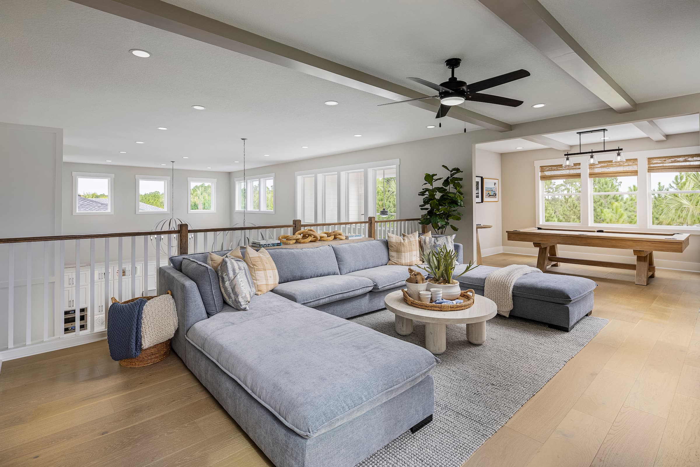 A spacious, modern living room with a large gray sectional sofa, round coffee table, pool table, ceiling fan, and natural light from multiple windows—perfect for enjoying life in one of the new homes in Wildlight FL.
