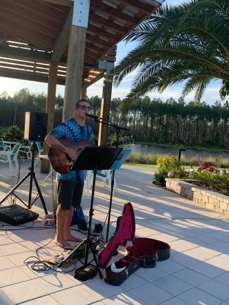 A person wearing sunglasses and a tie-dye shirt plays an acoustic guitar and sings into a microphone outdoors near a palm tree, with an open guitar case on the ground in the Wildlight master planned community.