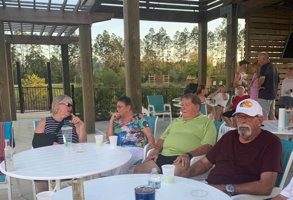 Four older adults sit around a white table outdoors, talking, in the heart of the Wildlight master planned community. Other people mingle under a wooden pergola, with trees and a fence providing a scenic backdrop.