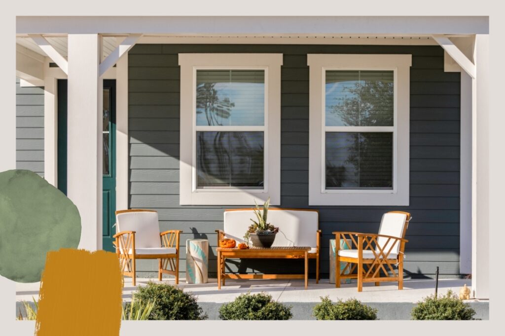 A front porch in the Wildlight master planned community features two windows, outdoor wooden furniture with white cushions, a small table with a plant, and shrubs in front—perfect for enjoying homes in Nassau County Florida.