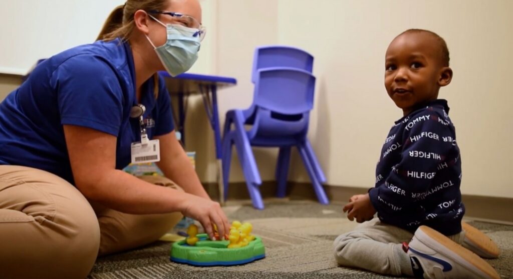 A healthcare worker wearing a mask plays a tabletop game with a young child in a cheerful room with blue chairs, reflecting the caring community spirit found in Wildlight Florida and its welcoming new homes in Wildlight FL.