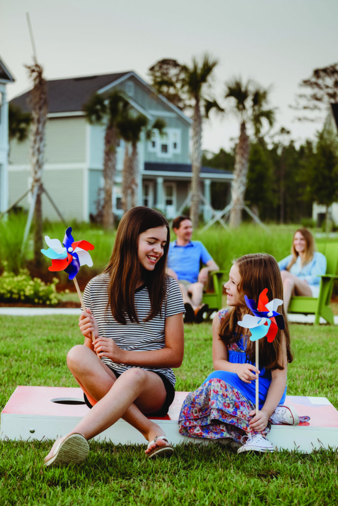 Two girls sit on the grass holding pinwheels and smiling at each other, while two adults relax nearby in front of new homes in Wildlight FL and swaying palm trees.
