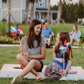 Two girls sit on the grass holding colorful pinwheels, smiling at each other, with two adults nearby—capturing a joyful moment in the welcoming Wildlight Yulee Florida community.