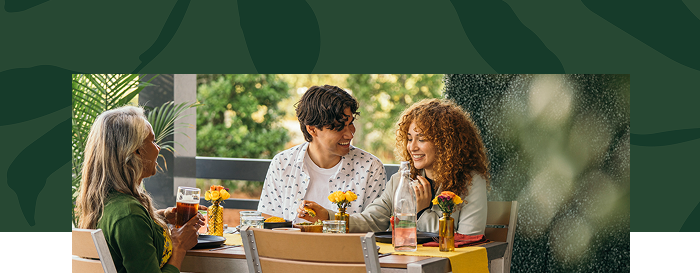 Three people sit at an outdoor table, enjoying drinks and conversation, with yellow flowers as table decorations and greenery in the background.