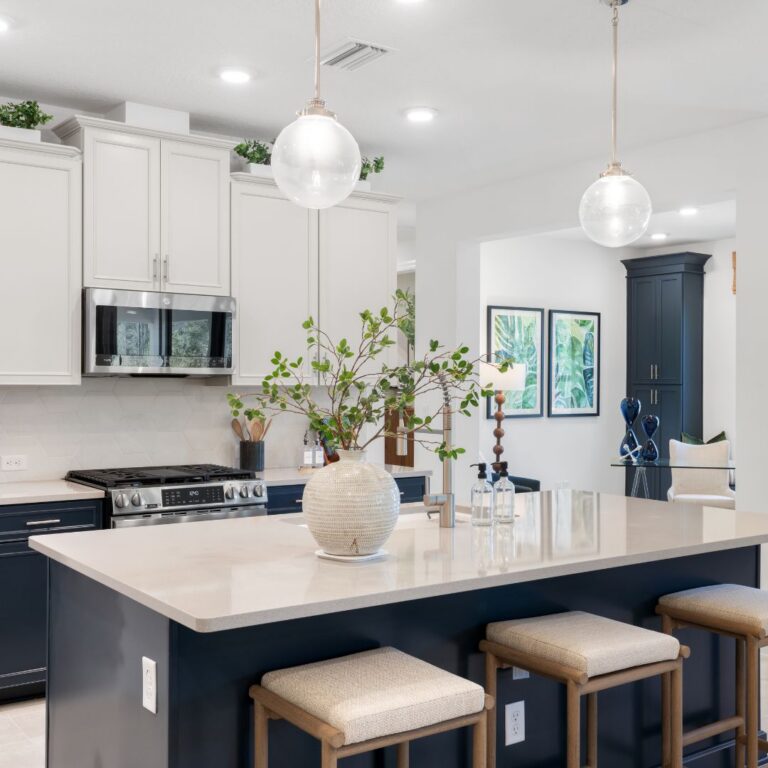 Modern kitchen with a large island, three stools, a vase with greenery, pendant lights, white cabinets, and stainless steel appliances.