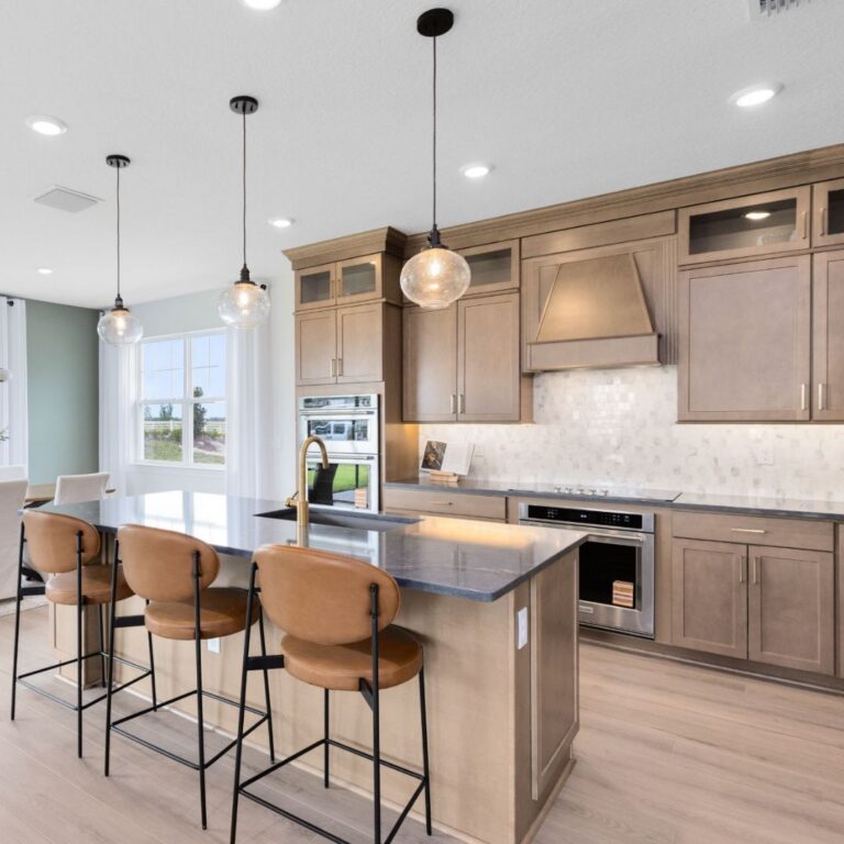 Modern kitchen with light wood cabinets, an island with three brown chairs, pendant lights, built-in oven, and large windows providing natural light.