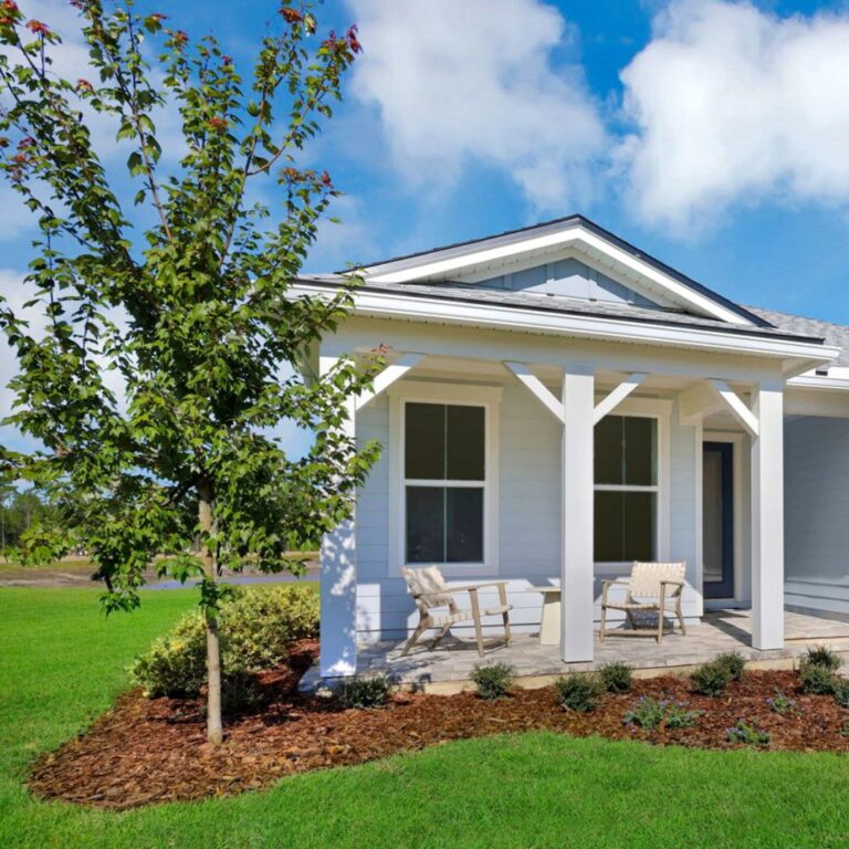 Single-story house with a covered front porch, two chairs, a small table, a young tree, and neatly landscaped yard under a blue sky with clouds.
