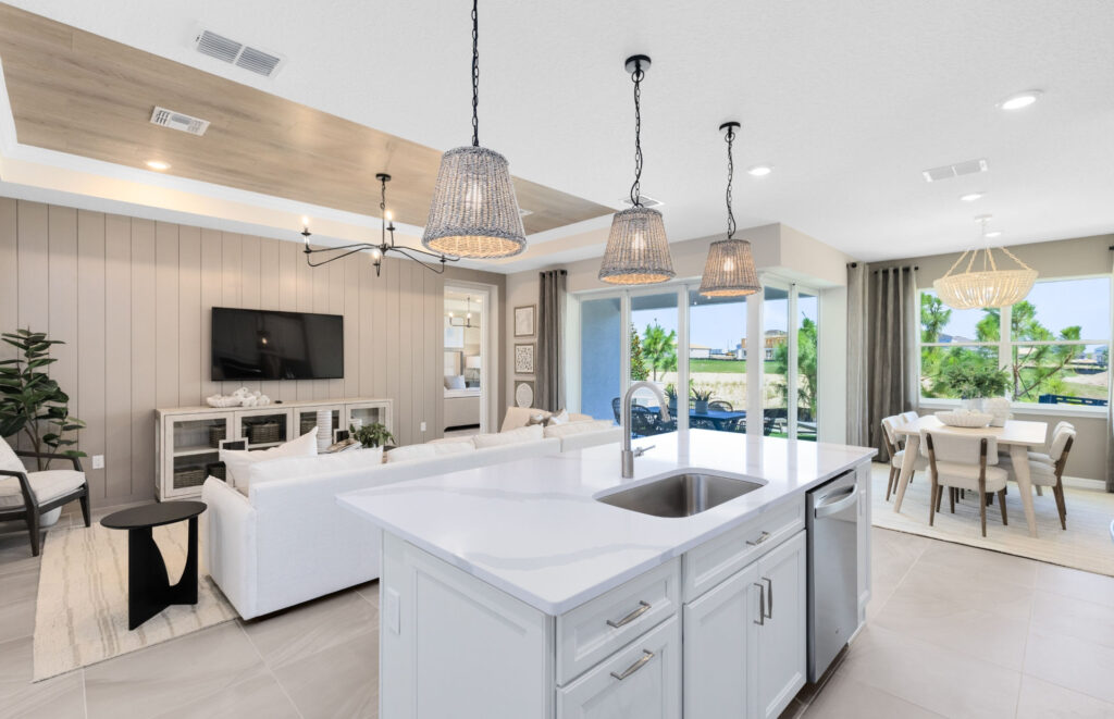 Modern open-concept kitchen and living area with white cabinetry, island sink, pendant lights, dining table, large windows, and a wall-mounted TV.